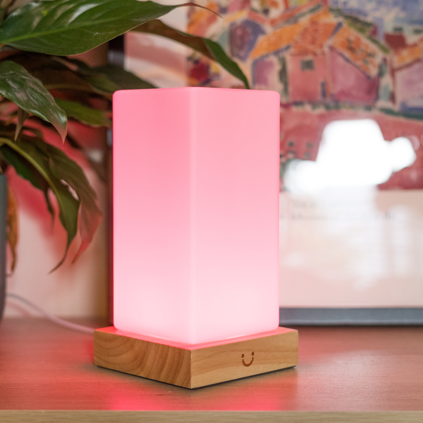 Pink tower lamp with wooden base glowing softly next to green plant on a desk.