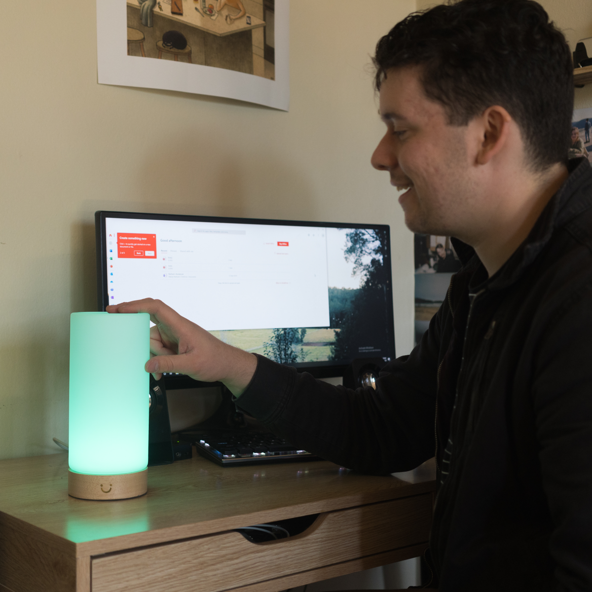 A man reaches toward a glowing mint-green cylinder lamp on his desk beside a computer monitor.