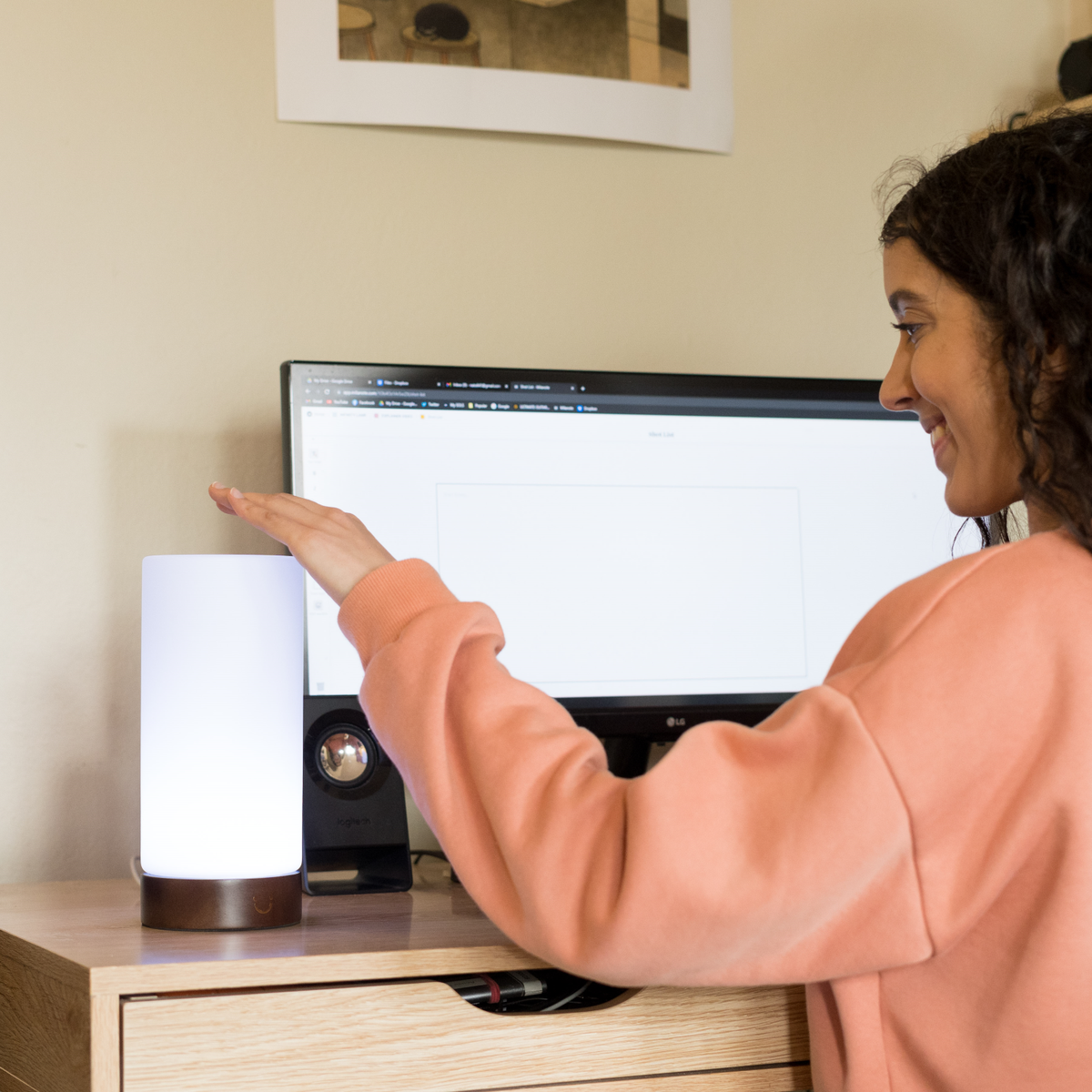 Woman gestures towards a white cylindrical lamp on her desk beside a monitor.